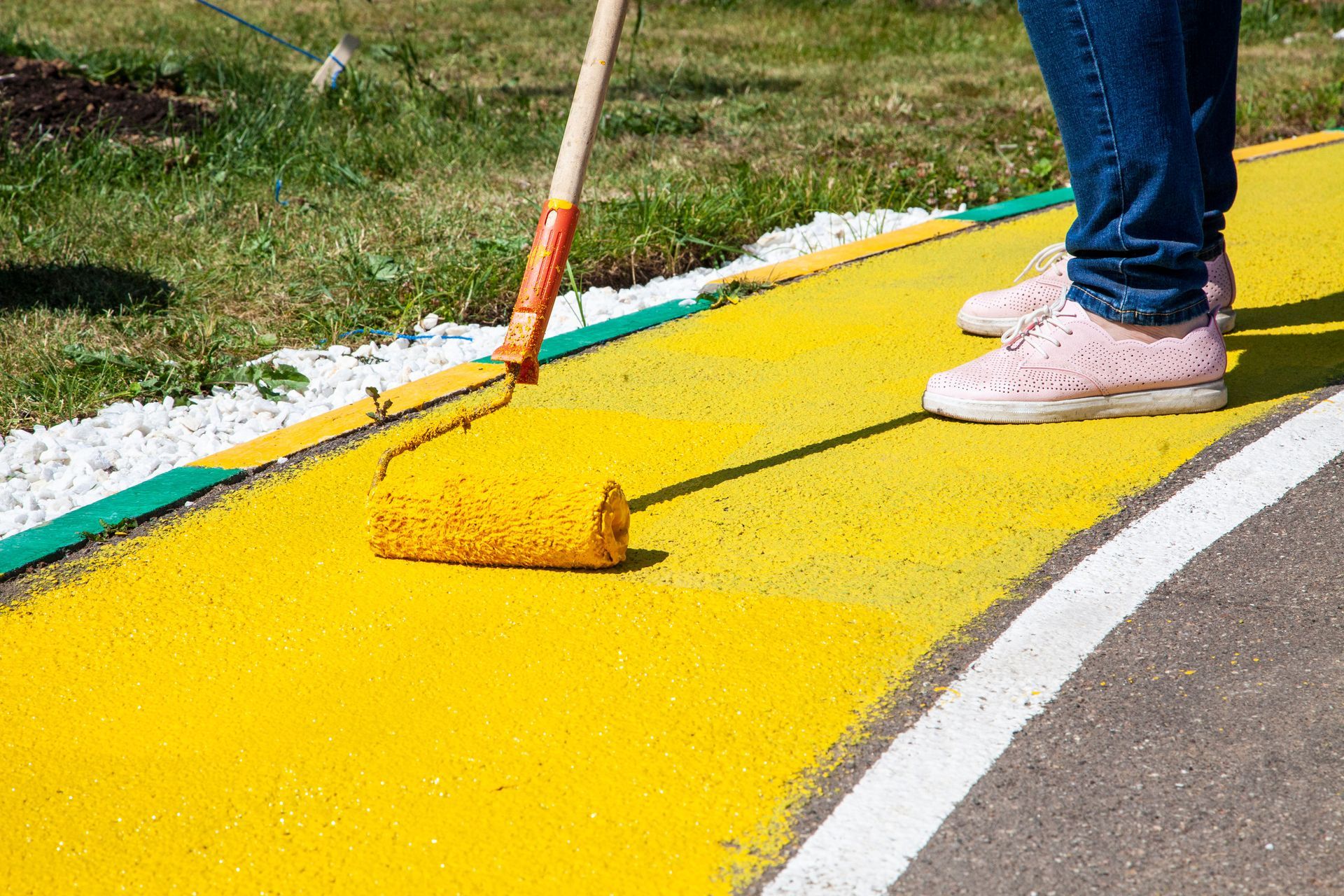 Persona con jeans y zapatillas rosas pintando un camino amarillo con un rodillo.