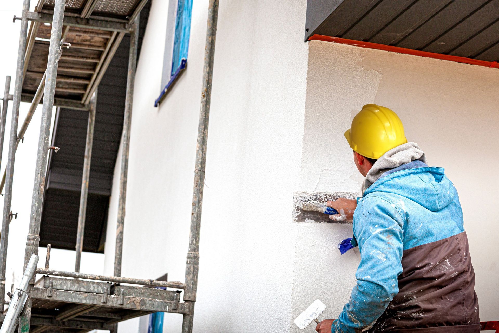 Obrero de la construcción con casco amarillo aplicando estuco blanco a la pared exterior de un edificio, con andamios al fondo.