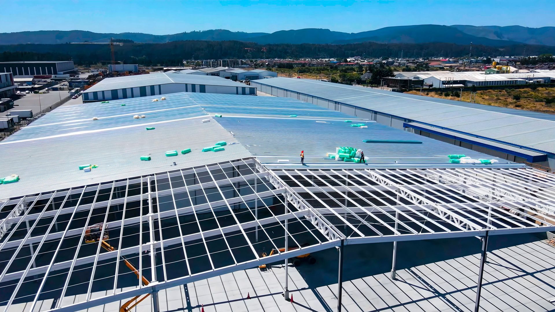 Vista aérea de un gran almacén en construcción, con techo metálico parcialmente terminado. Cielo azul y montañas al fondo.