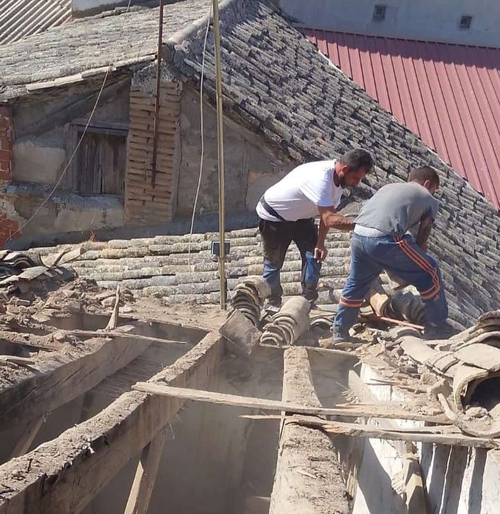 Dos pintores, uno de pie y otro arrodillado, trabajan en el exterior blanco de un edificio. El edificio tiene detalles en rojo y un techo gris.