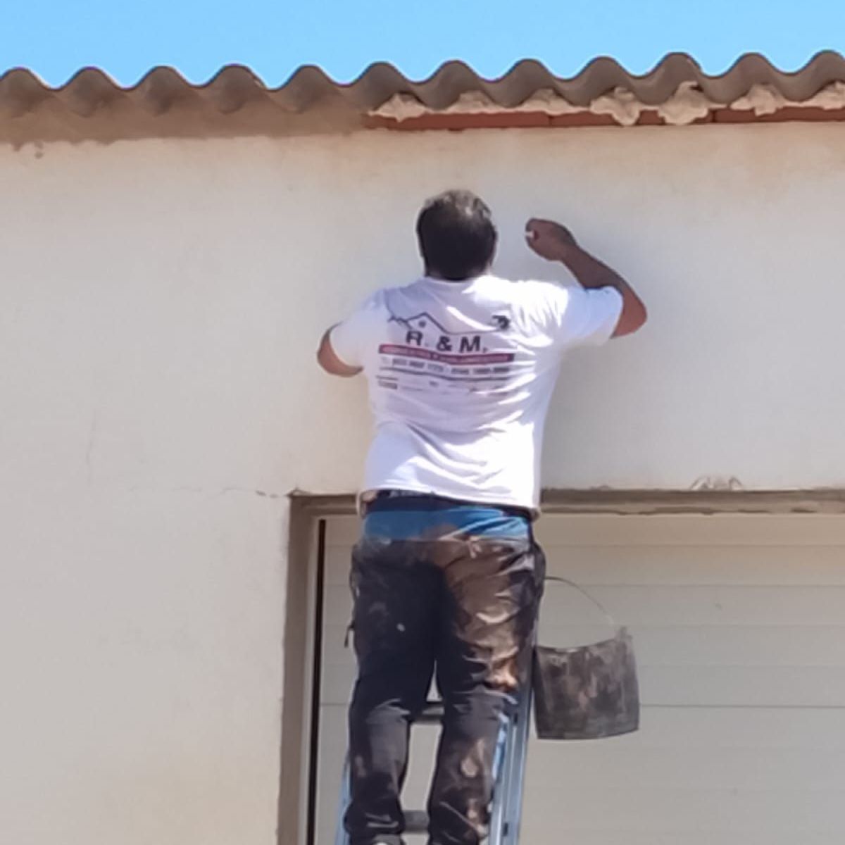 Dos pintores en un andamio aplican estuco al exterior de un edificio, con una pared roja y un cielo azul visibles.