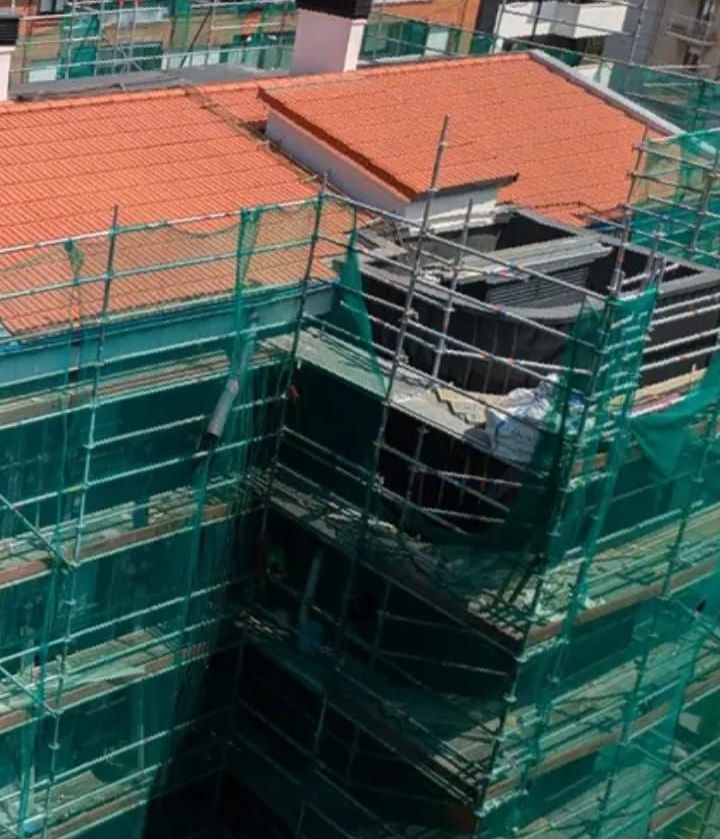 Persona pintando una pared rosa. Lleva guantes azules y usa una brocha y una bandeja de pintura. También hay una escalera, rodillos y otros materiales de pintura.