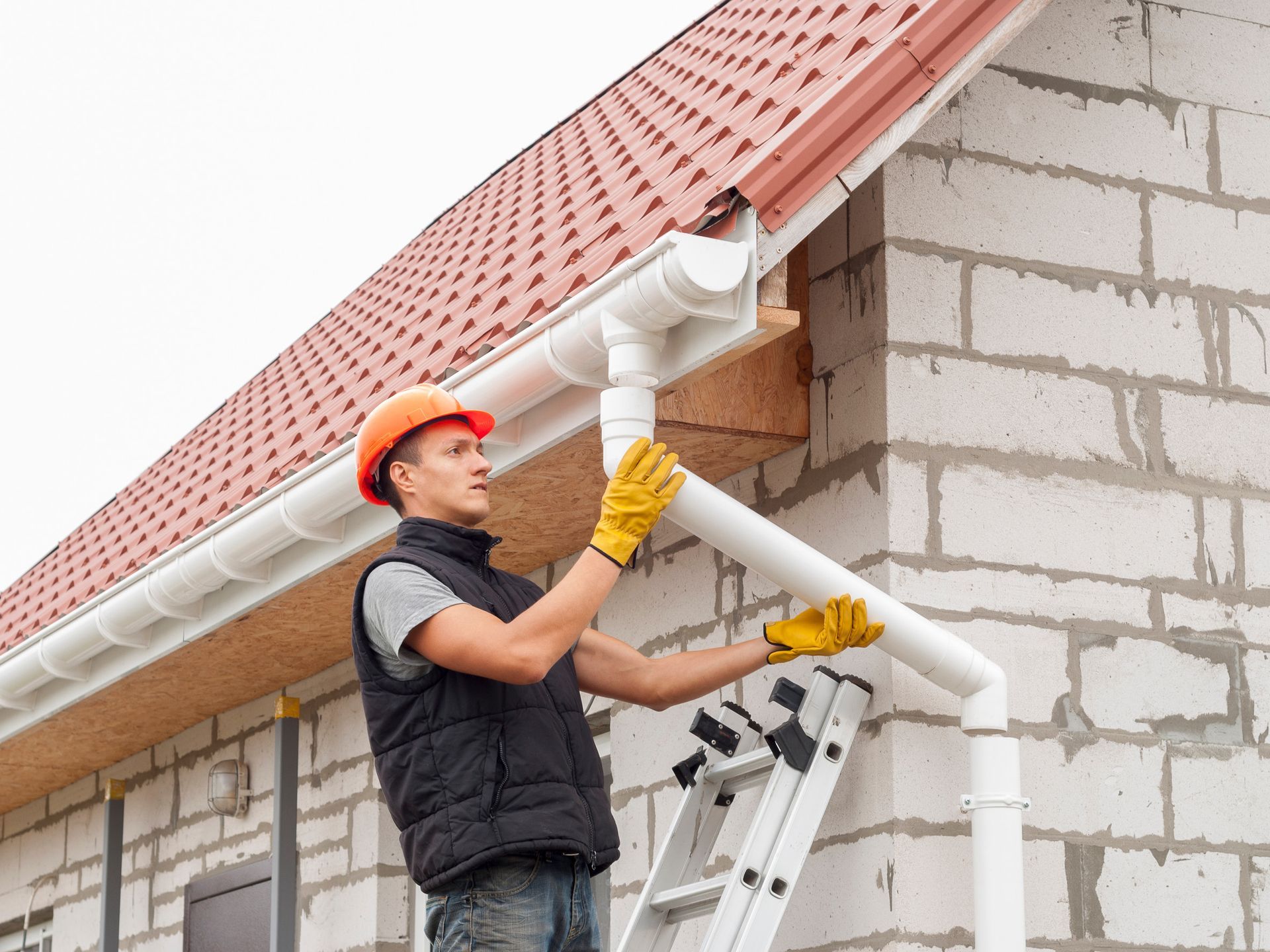 Hombre con casco y guantes instalando una canaleta blanca en un edificio de ladrillos.