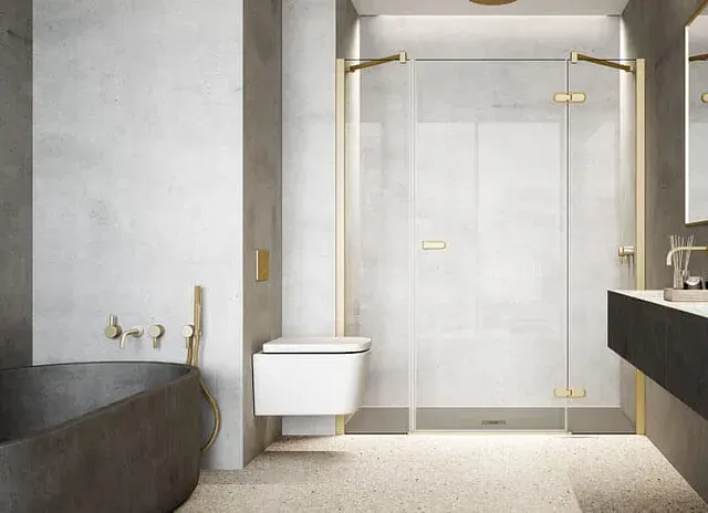 Baño moderno con paredes de hormigón gris, detalles dorados, ducha de cristal, bañera negra y inodoro flotante.