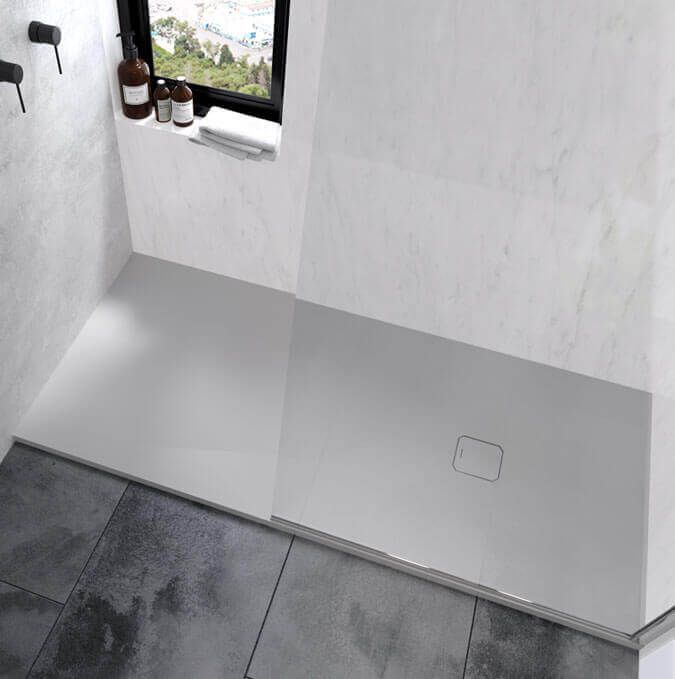 Plato de ducha gris oscuro con desagüe, junto a un panel de vidrio. Azulejos grises y blancos al fondo.
