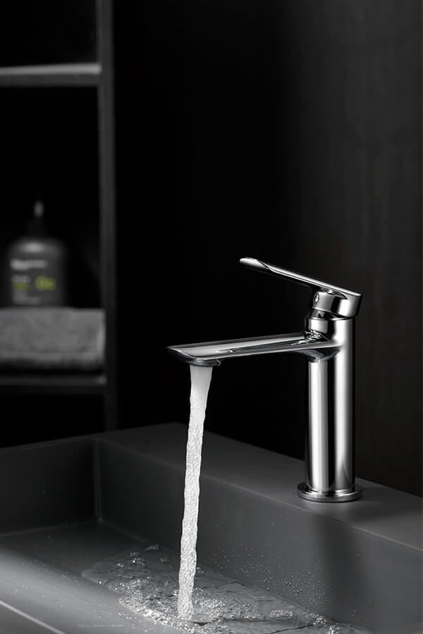 Grifo cromado con agua saliendo de un lavabo gris oscuro. Fondo: pared negra y estantes.