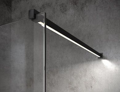 Luminaria lineal negra montada en una pared de hormigón gris, que ilumina un panel de ducha de vidrio.