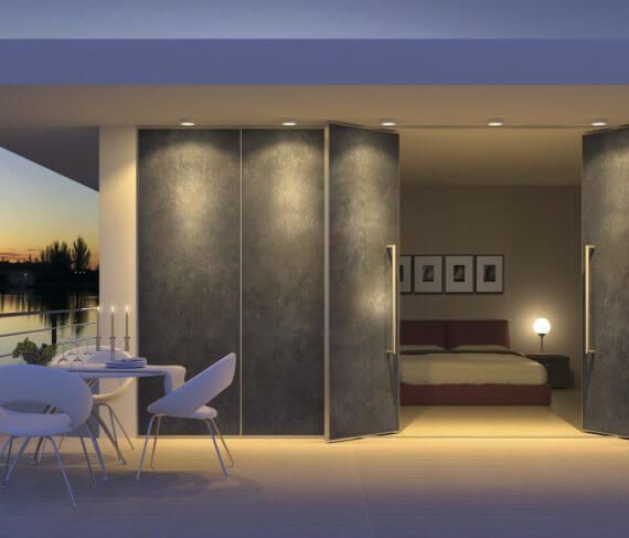 Las puertas abiertas revelan un dormitorio desde un balcón con mesa y sillas. Atardecer sobre el agua.