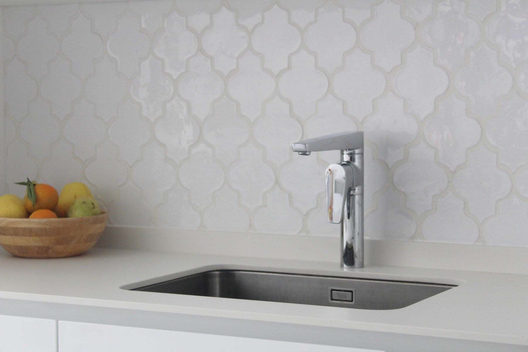 Fregadero de cocina de acero inoxidable y grifo sobre encimera blanca con protector contra salpicaduras de azulejos decorativos.
