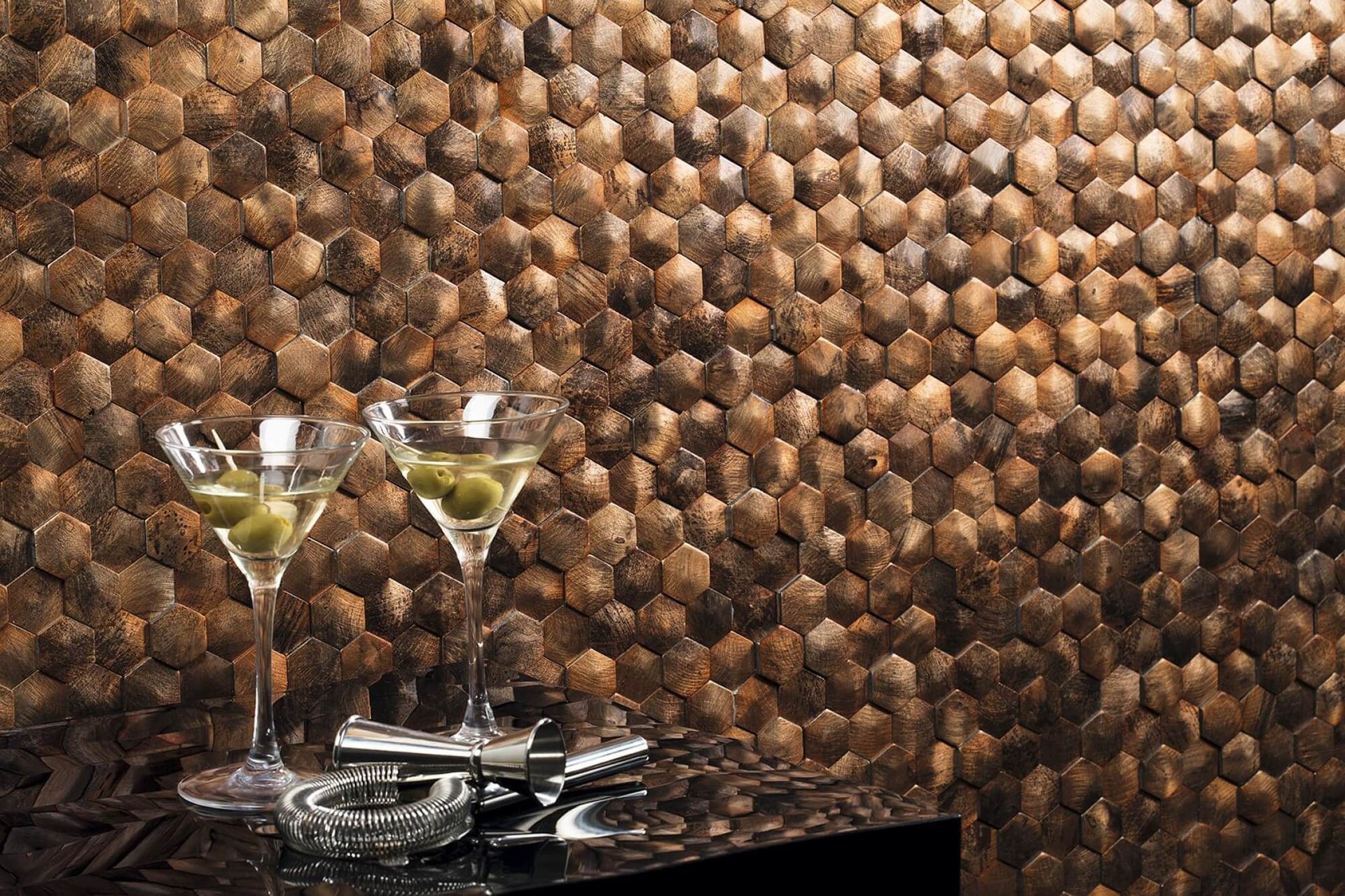 Dos martinis con aceitunas sobre una superficie negra contra una pared de azulejos marrones con diseño en forma de panal.