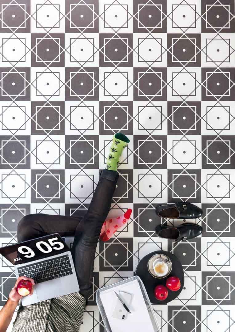 Persona sentada en el suelo con un portátil, un libro y bebidas. Azulejos geométricos en blanco y negro; un calcetín verde.