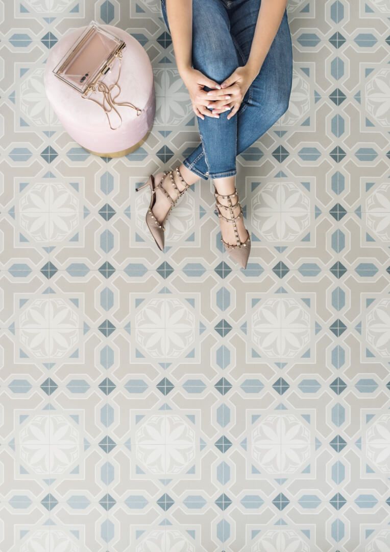 Persona sentada en un piso de baldosas estampadas con un taburete rosa, vistiendo jeans y tacones.