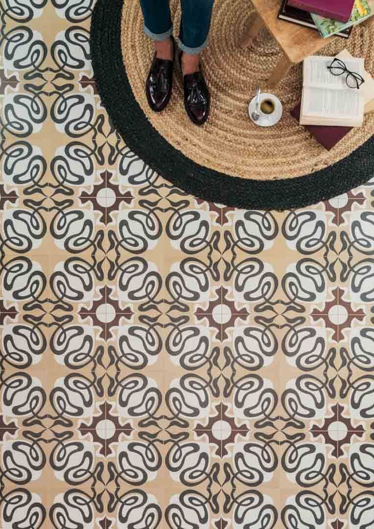 Persona de pie junto a una alfombra con café y libros sobre un piso de baldosas estampadas.