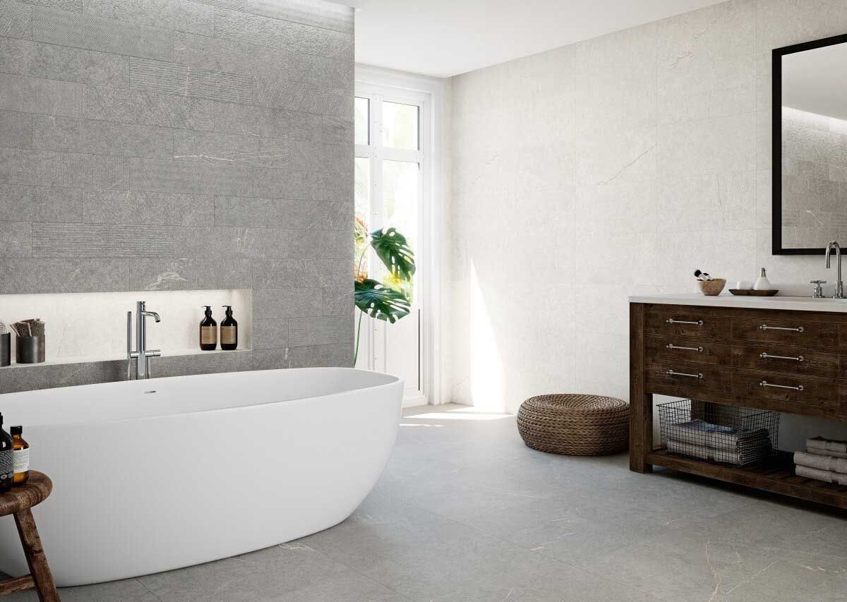 Baño moderno con bañera blanca, pared de piedra gris, tocador de madera y ventana grande.