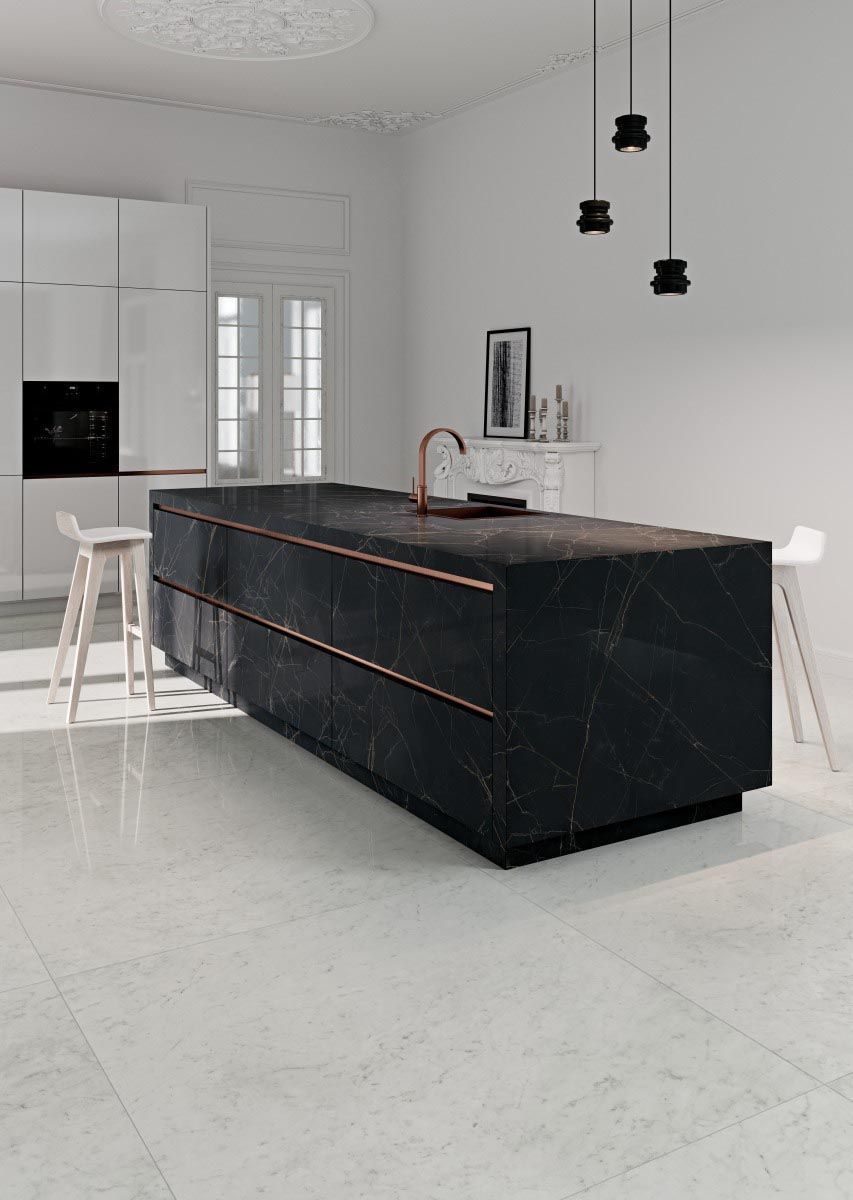 Isla de cocina de mármol negro con detalles en cobre, gabinetes blancos y pisos claros.