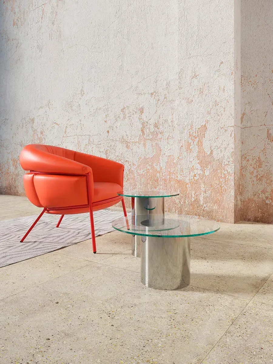 Sillón naranja y dos mesas con tapa de cristal sobre un suelo de color claro, contra una pared texturizada.