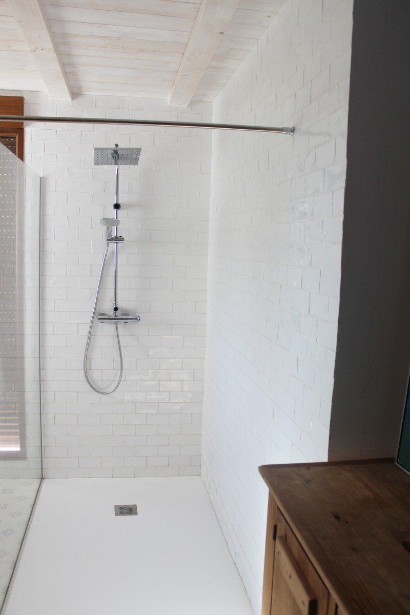 Ducha de azulejos blancos con cabezal de lluvia y ducha de mano, contra una pared blanca texturizada.