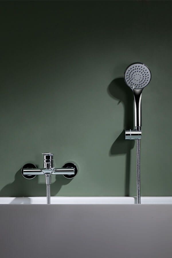 Grifo de ducha cromado y cabezal de ducha de mano contra una pared verde apagado, montado encima de una bañera blanca.