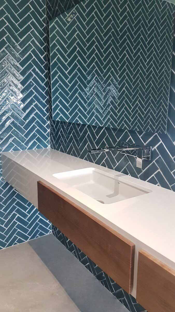 Baño con salpicadero de azulejos en espiga azul, encimera blanca y mueble de madera.