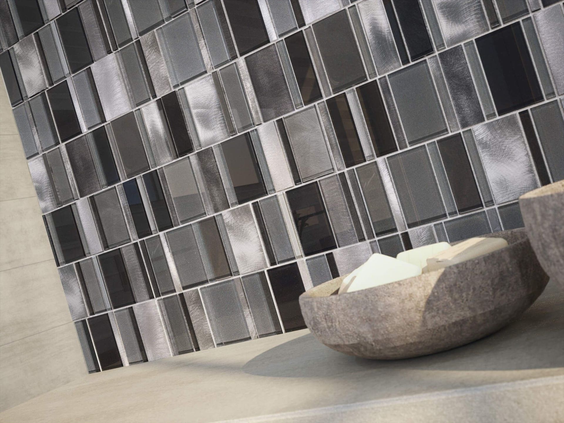 Pared de mosaico rectangular gris y negro con un cuenco de piedra lleno de objetos blancos.