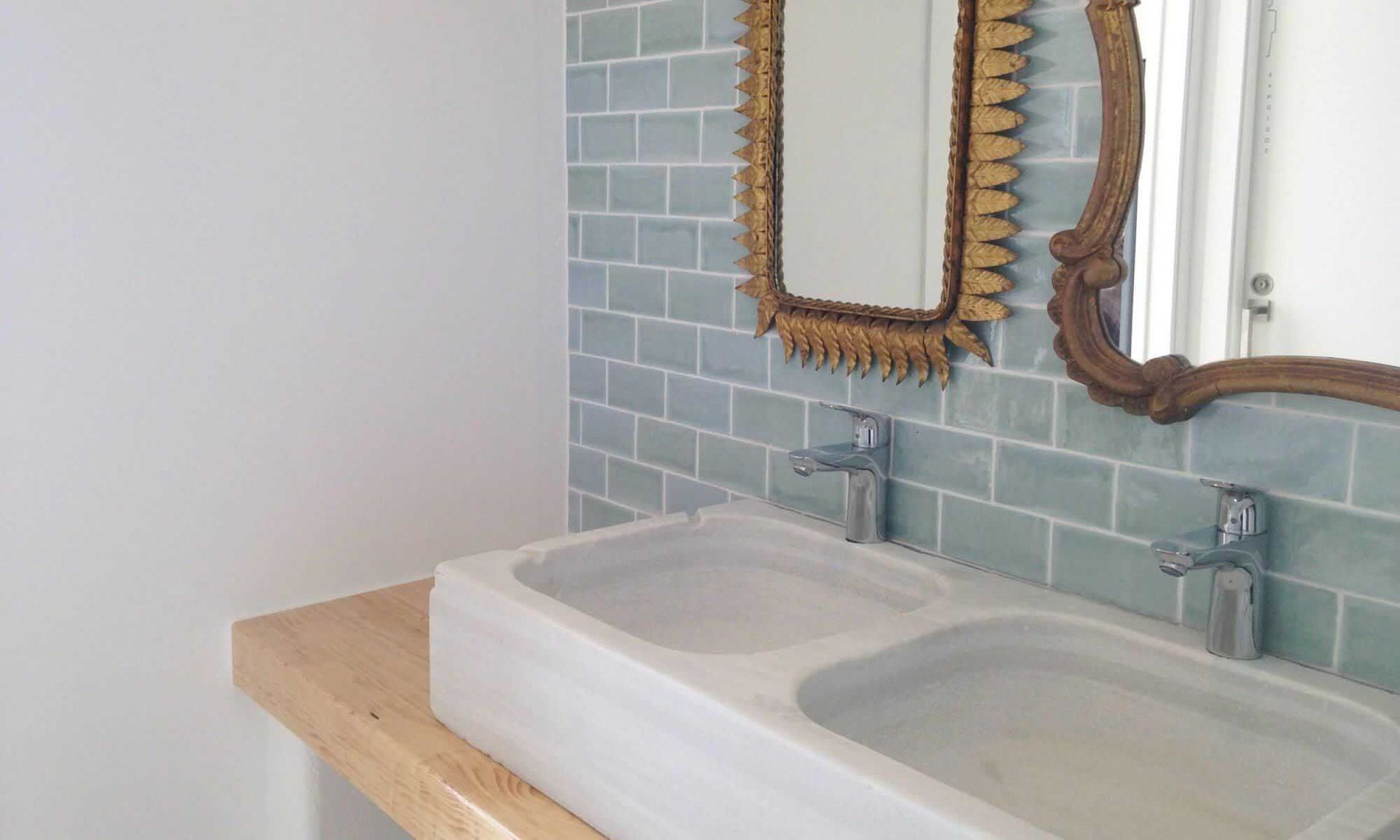 Lavabos dobles en el baño con pared de azulejos azules, dos espejos ornamentados y un estante de madera.