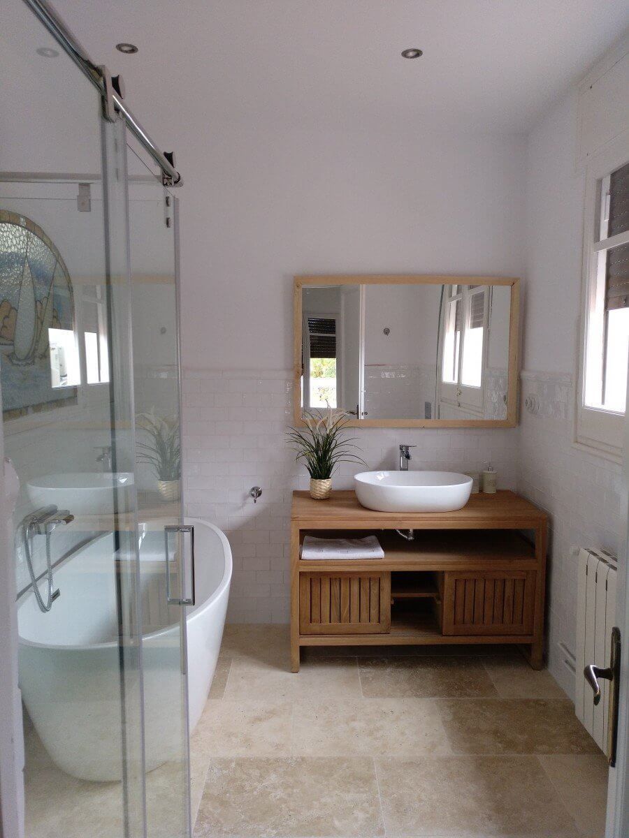 Baño luminoso con bañera profunda blanca, tocador de madera con lavabo de pedestal y puerta de ducha corrediza de vidrio.