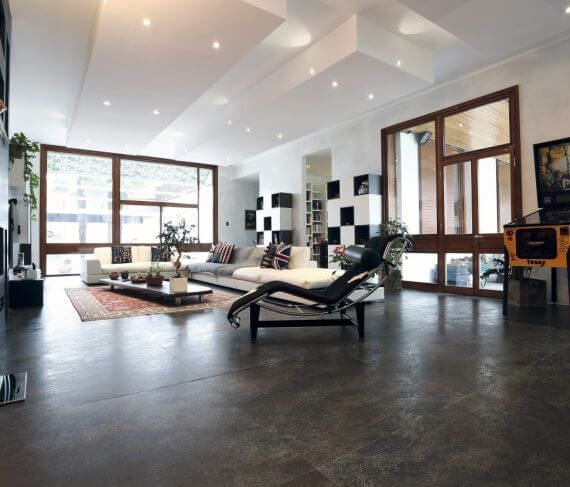 Amplia sala de estar moderna con grandes ventanales, pisos oscuros y decoración minimalista; un diván negro se encuentra en primer plano.