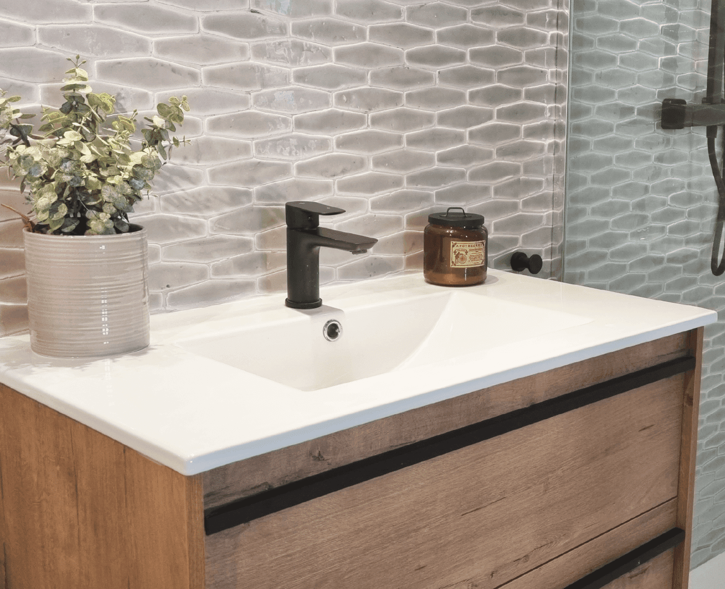 Tocador de baño con encimera blanca, mueble de madera marrón, grifo negro y salpicadero de azulejos grises estampados.