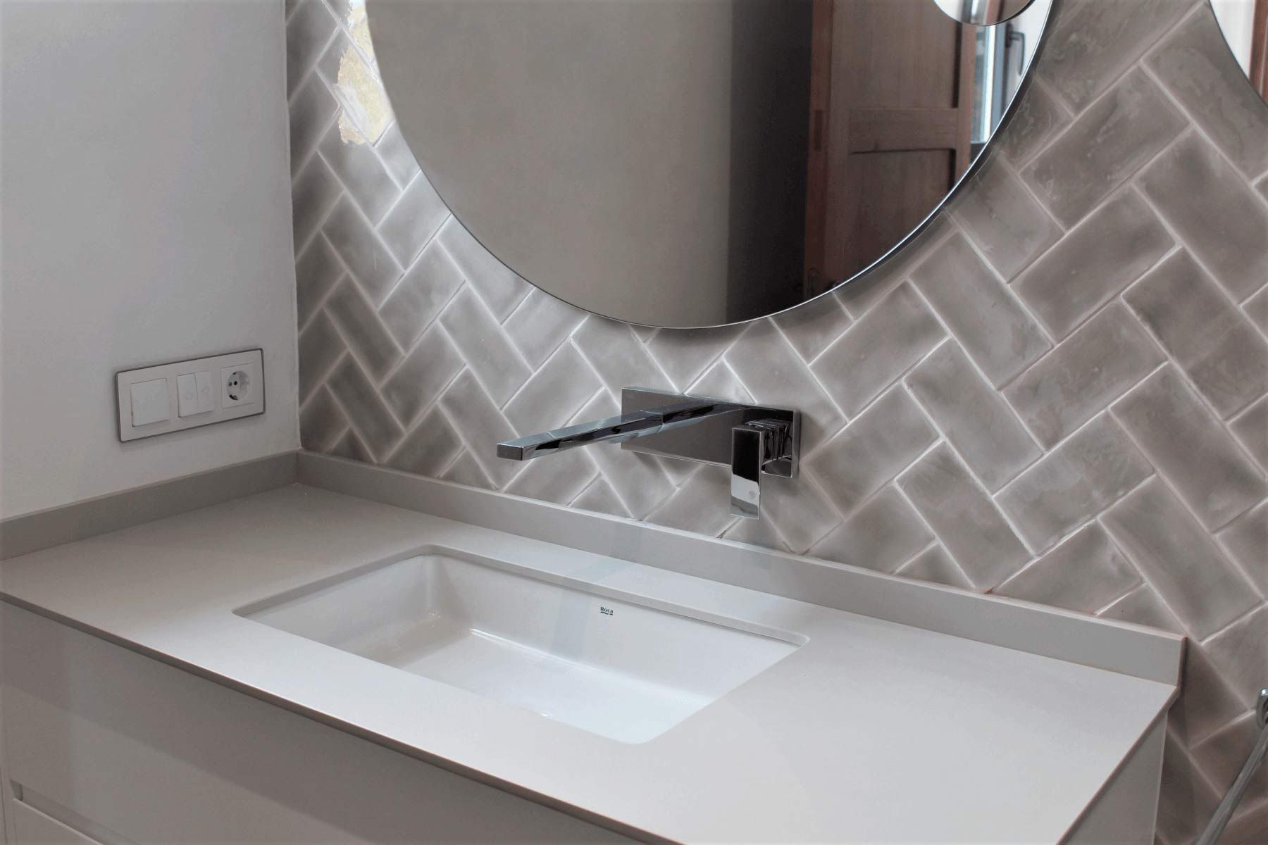 Baño moderno con lavabo rectangular, grifo, espejo redondo y pared decorativa con azulejos grises.