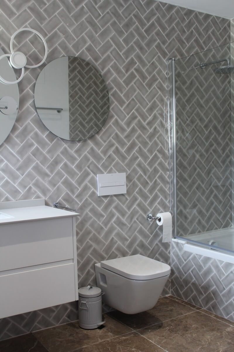 Baño con azulejos grises en espiga, inodoro blanco, tocador y espejos. Incluye ducha y papelera.
