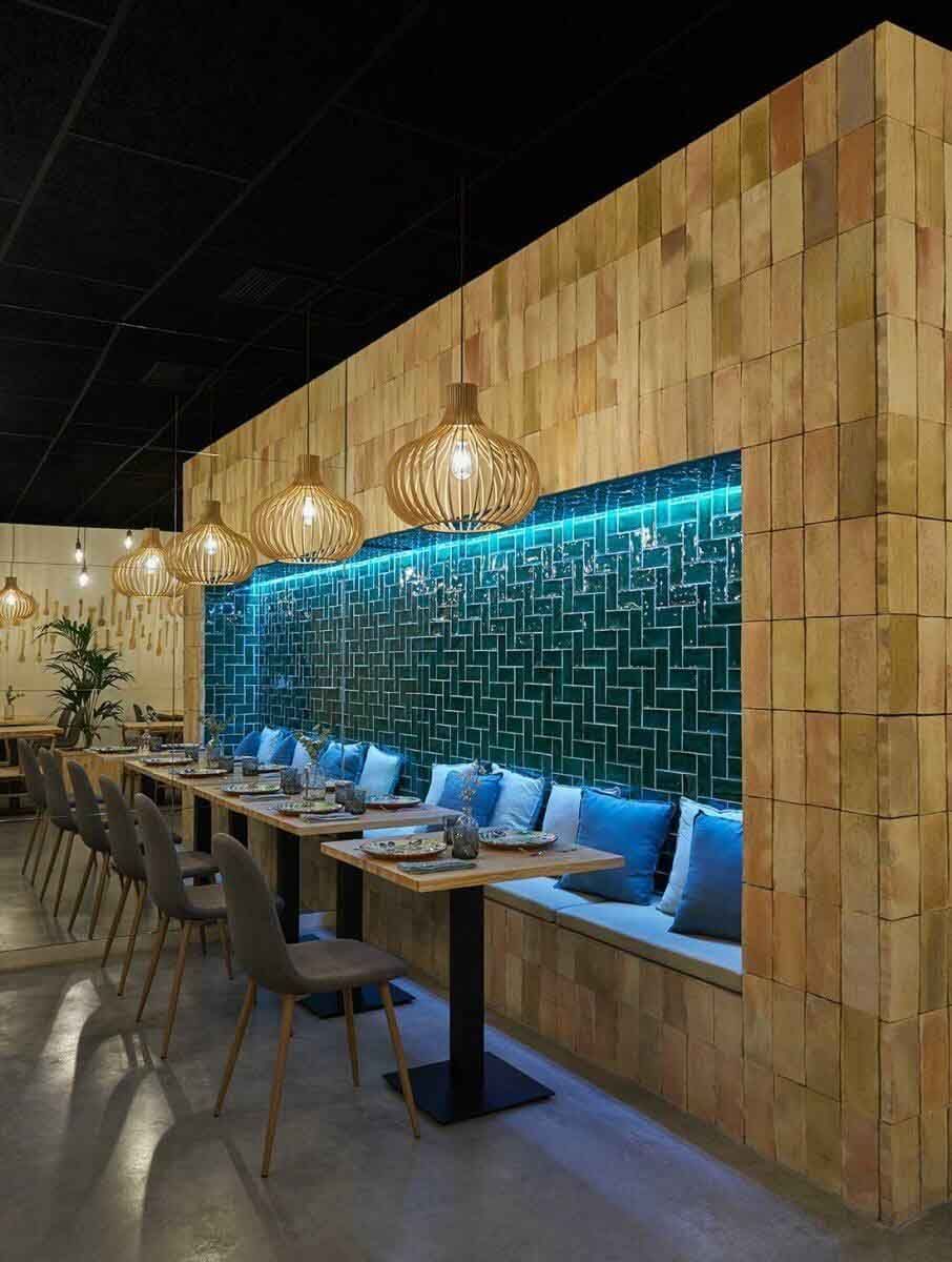 Interior de restaurante con paredes de paneles de madera, azulejos turquesas y luces colgantes sobre una cabina con almohadas azules.