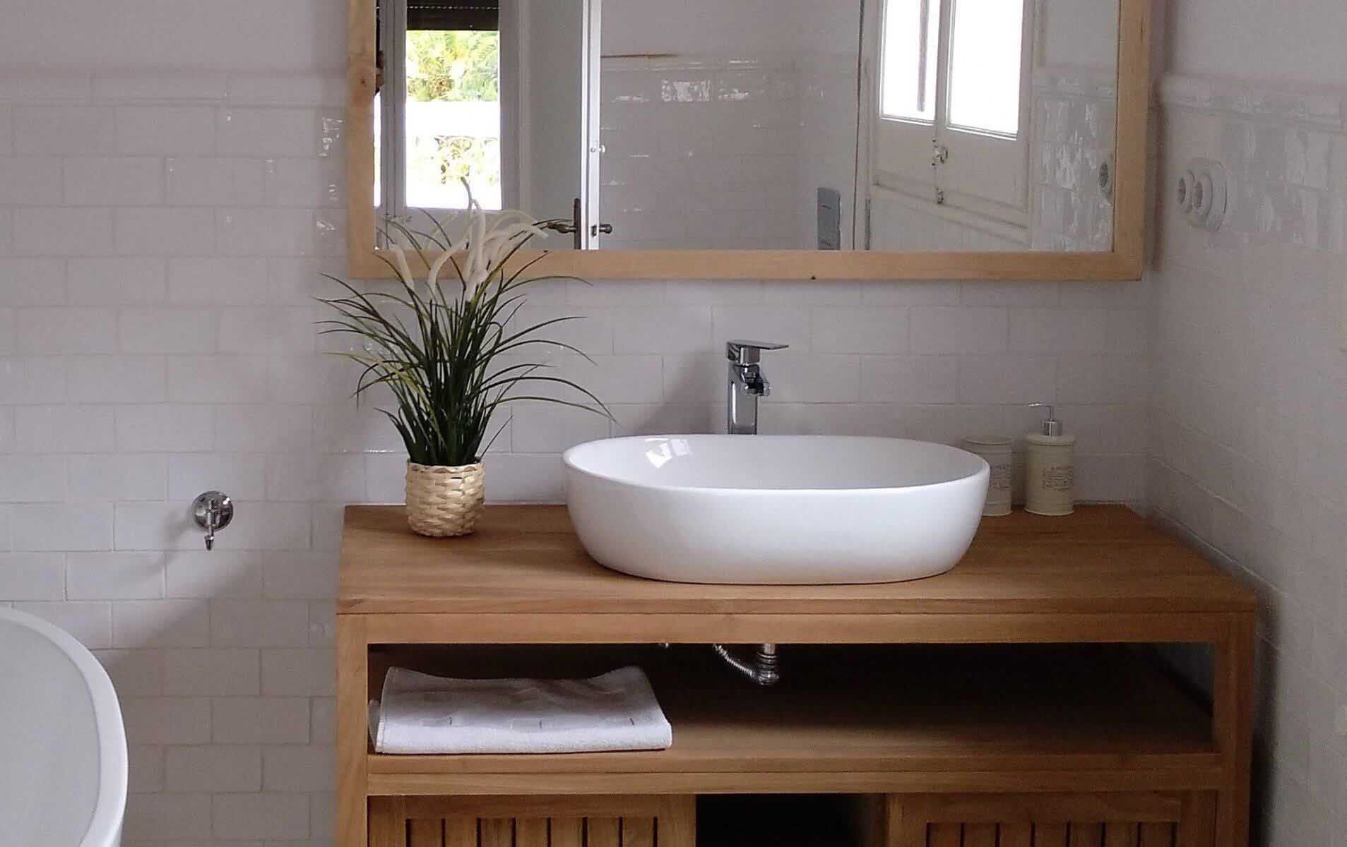 Baño con tocador de madera, lavabo blanco y espejo grande. Planta en una cesta. Paredes de azulejos blancos.