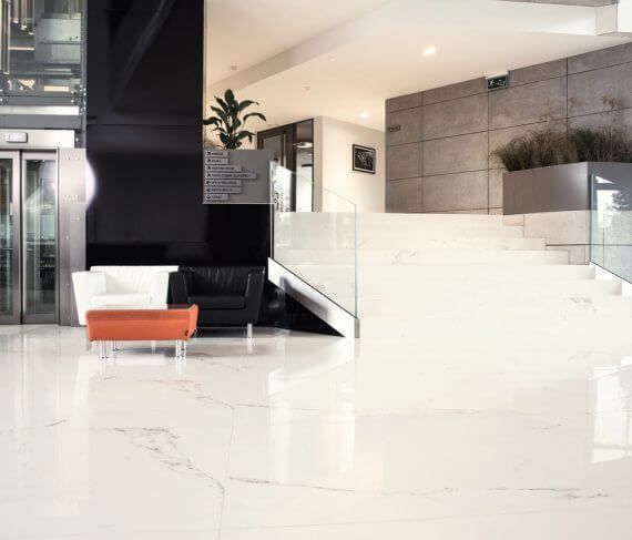 Moderno vestíbulo con suelo de mármol blanco, zona de estar, escalera y ascensor.