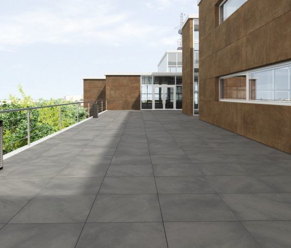 Amplio patio de baldosas grises junto a un edificio marrón con ventanas y barandilla de cristal. Árboles al fondo.