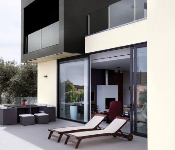 Exterior de casa moderna con patio y balcón. Fachada en negro y beige, puertas de cristal y sillones.