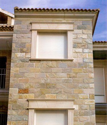 Fachada de edificio de piedra con dos ventanas con persianas blancas y molduras de piedra ornamentadas.