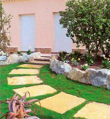 Camino de piedras cuadradas a través de césped verde hacia un edificio de color rosa claro con escalones y un jardín de rocas.