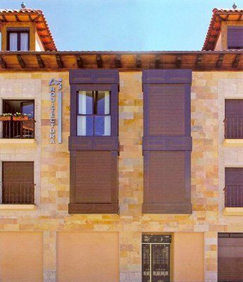 Fachada de edificio de piedra con contraventanas y puertas marrones, con un cartel que dice 