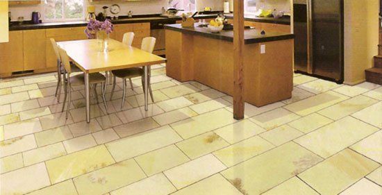 Cocina con piso de baldosas claras, gabinetes de madera, isla y mesa con sillas; flores en la mesa.