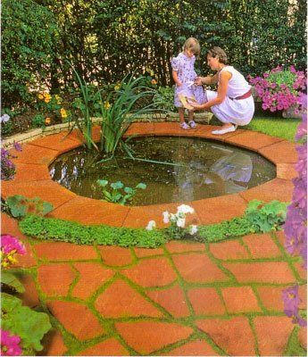 Mujer y niño junto a un estanque circular en un jardín, rodeado de ladrillos rojos y flores.