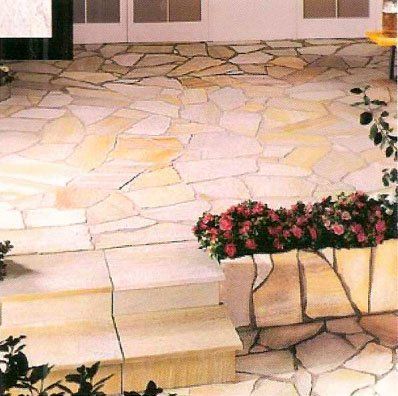 Patio de piedra beige con baldosas de forma irregular, escalones y flores rosas en macetas.