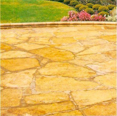 Patio de piedra amarilla con césped verde y flores rosas al fondo.