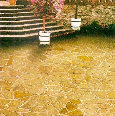 Patio de piedra con losas irregulares de color marrón amarillento, frente a escalones, flores en macetas y una fuente de agua oscura.