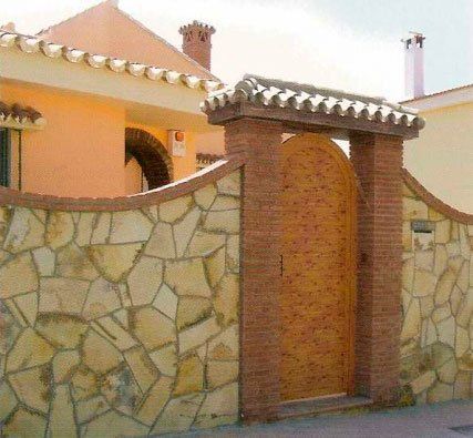 Muro de piedra con puerta de madera arqueada que conduce a una casa de color melocotón con techo de tejas.