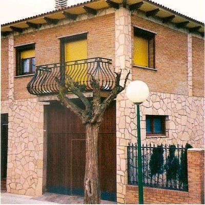 Edificio de ladrillo de dos plantas con detalles en piedra. Balcón con barandilla de hierro forjado. Árbol al frente.
