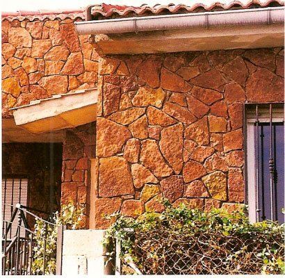 Exterior de un edificio con fachada de piedra roja, techo y ventana a la vista. Arbustos al frente.