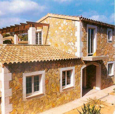 Casa de piedra de dos plantas con techo de tejas y ventanas con marcos blancos bajo un cielo soleado.