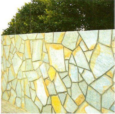 Muro de piedra con piedras de forma irregular de color verde claro y amarillo, rematado por follaje verde.