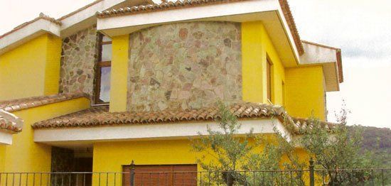 Una casa amarilla con detalles de piedra y techo de terracota, con algo de vegetación visible.