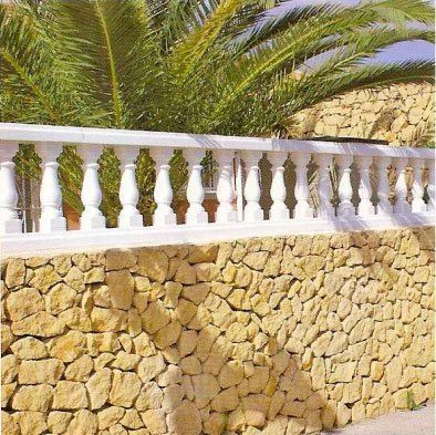 Muro de piedra rematado con balaustrada blanca, con hojas de palmera al fondo.